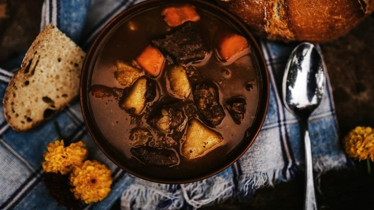 En gryta med köttbitar, potatis och morötter i en brun skål bredvid bröd och en sked.