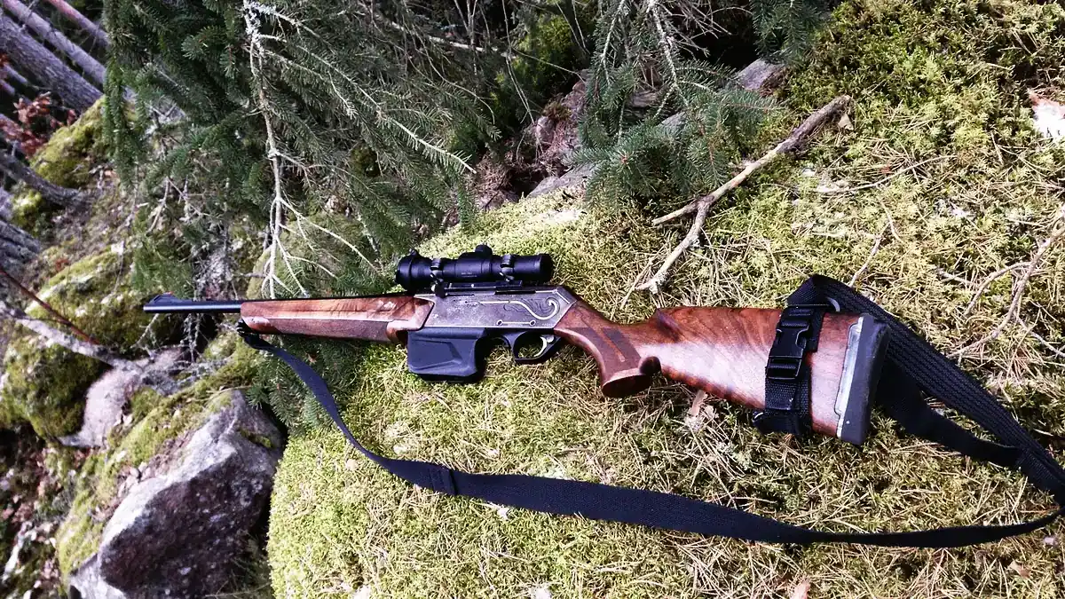 Ett kulgevär med kikarsikte och bärrem ligger på mossig mark i en granskog.