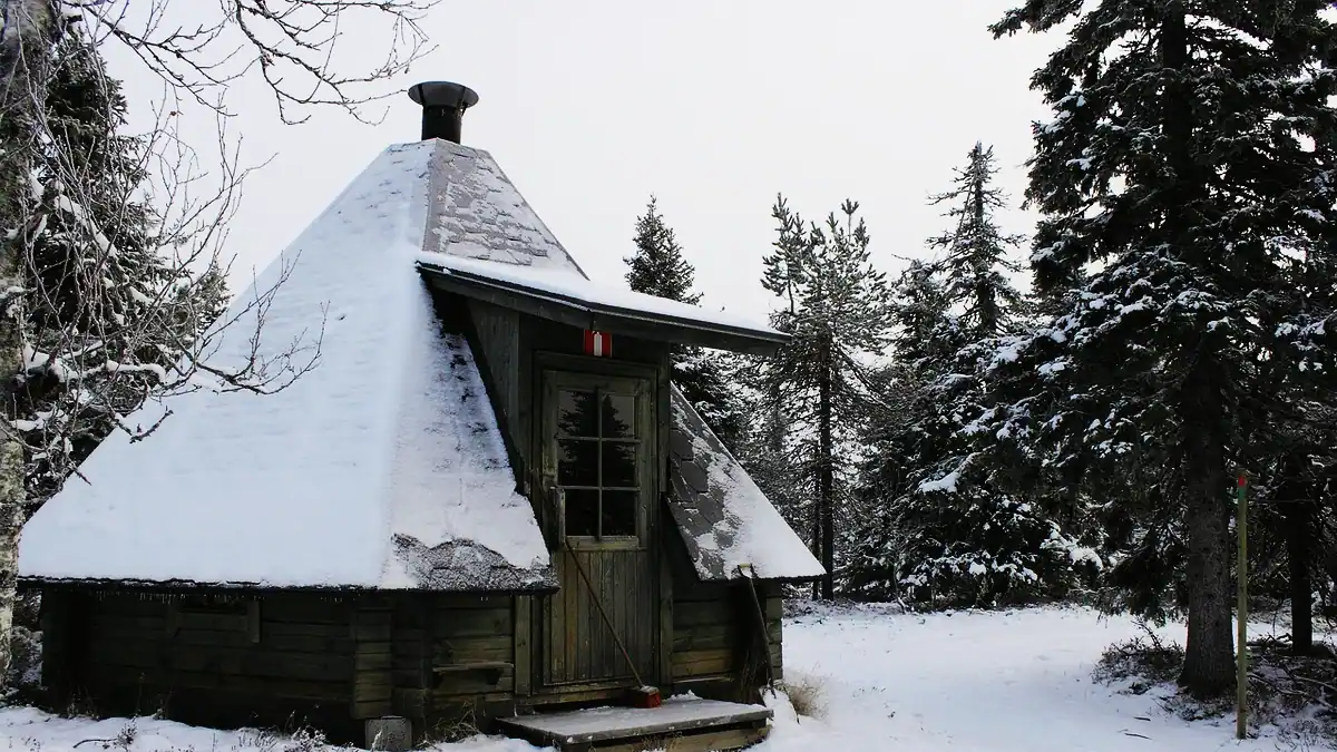En åttakantig timrad kåta med dörr, fönster och skorsten i snöig granskog.